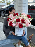 A person sitting on a curb holding a round white box of flowers with a variety of red, pink, and white flowers.