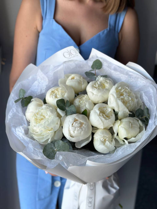 A person holding a bouquet of white peonies and greenery wrapped in paper.